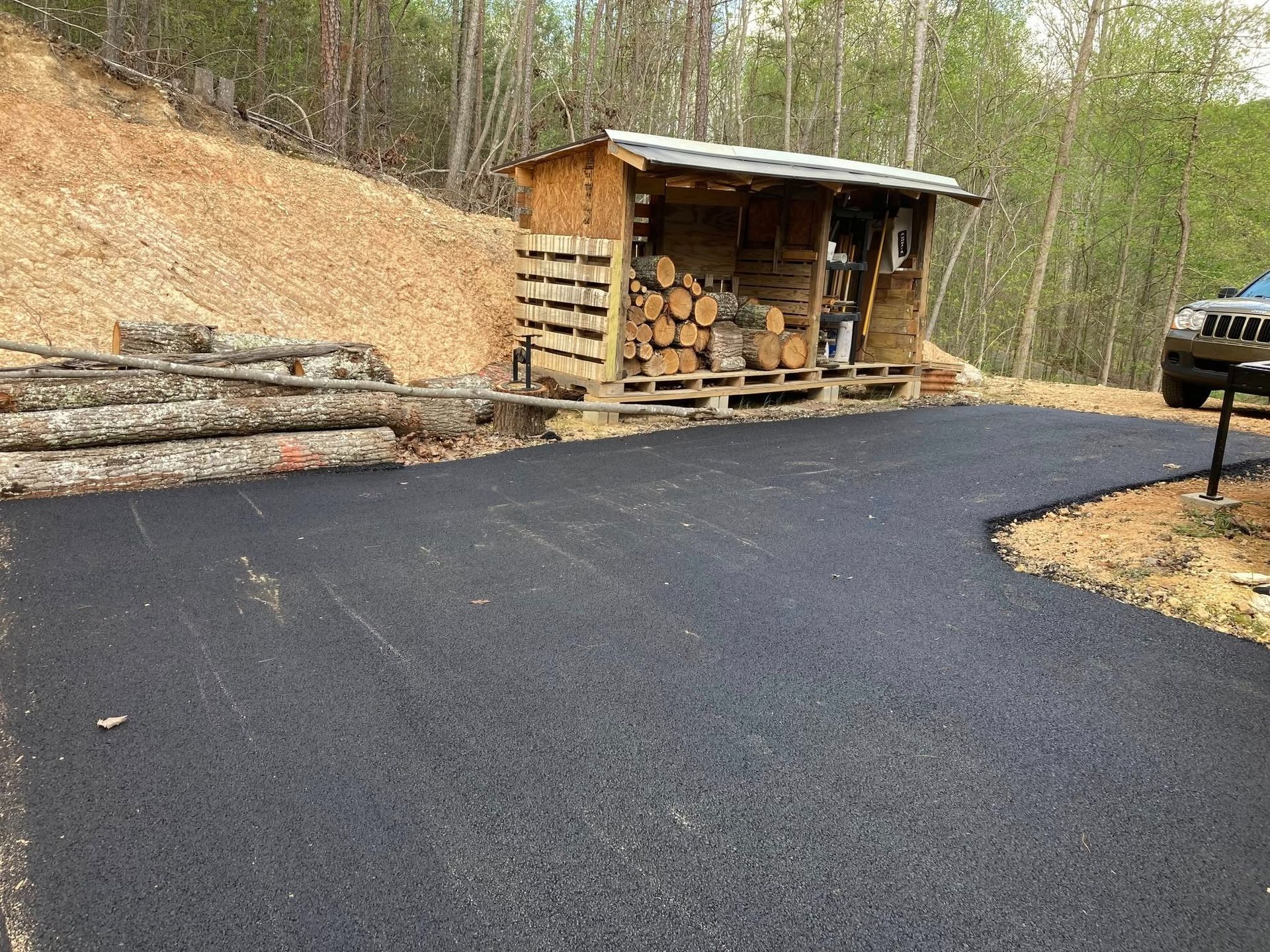A newly paved black asphalt driveway sits next to a wooden shed stacked with firewood, near a hill and a parked vehicle.