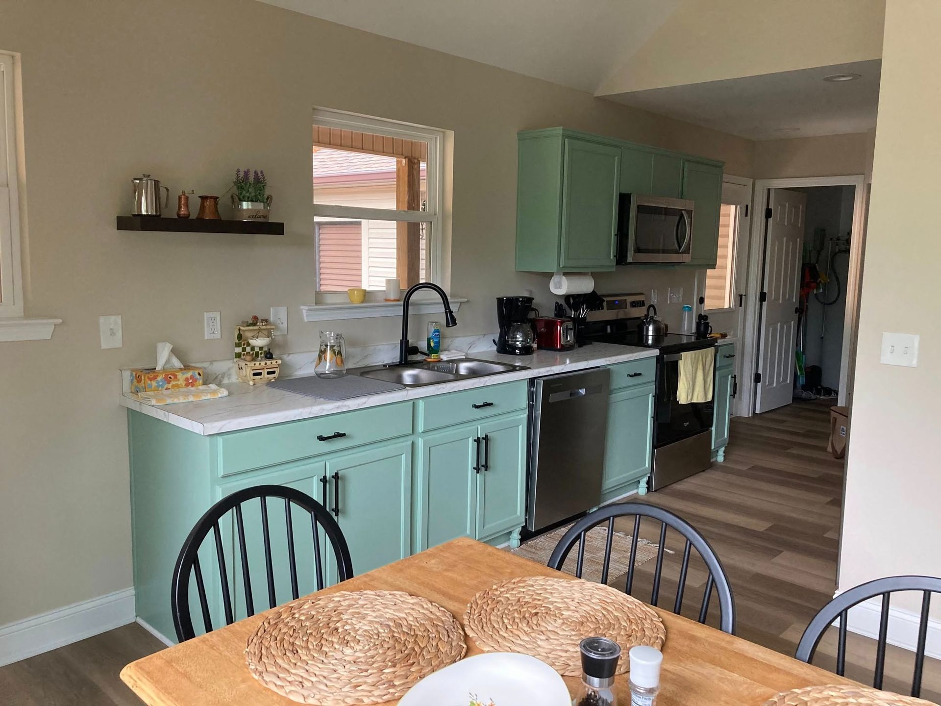 A bright kitchen with sage green cabinets, stainless steel appliances, a wooden dining table, and a small floating shelf.