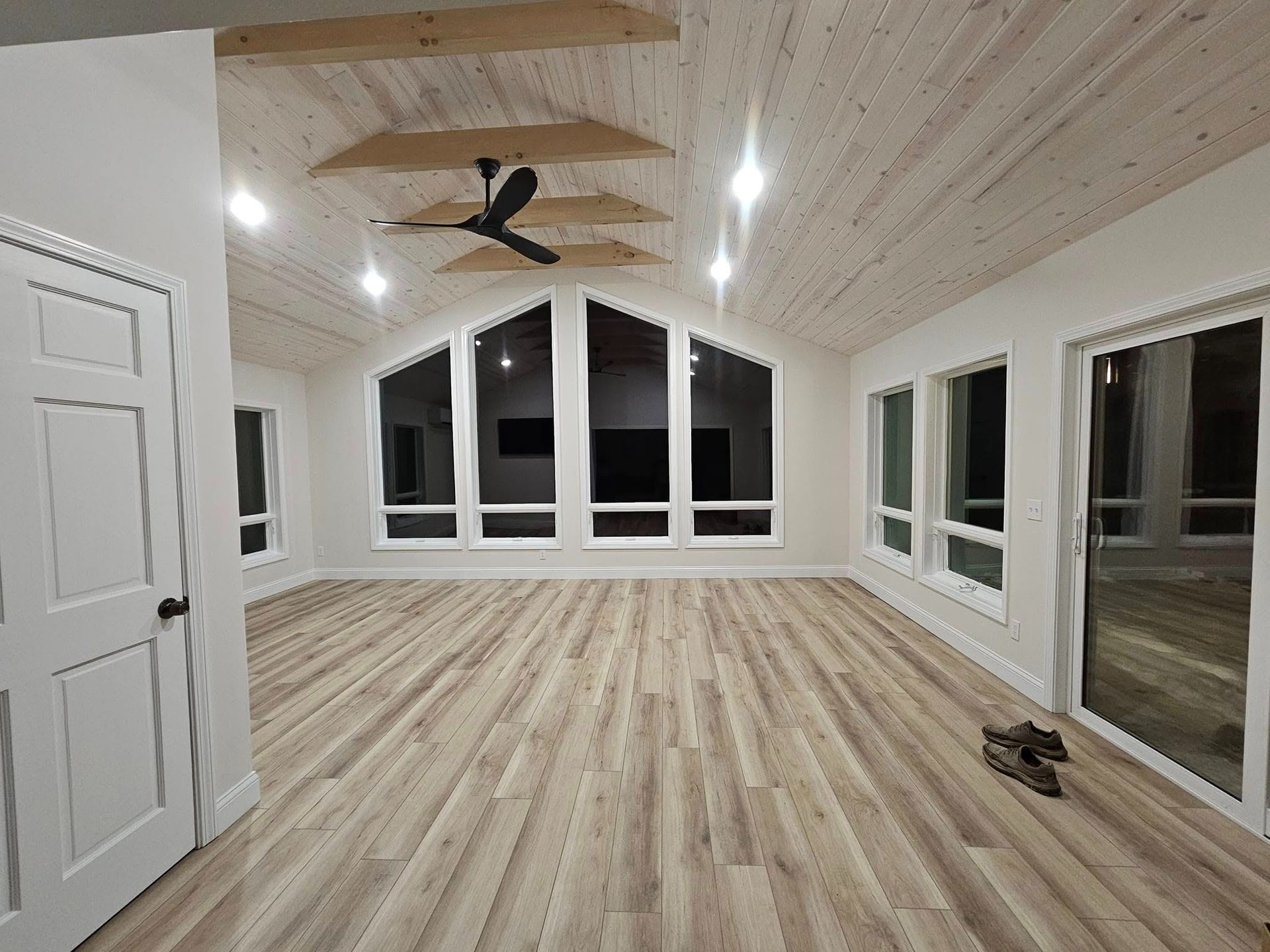 A spacious, empty room with light-colored wood-look flooring, white walls, and a vaulted, wood-paneled ceiling with a fan.