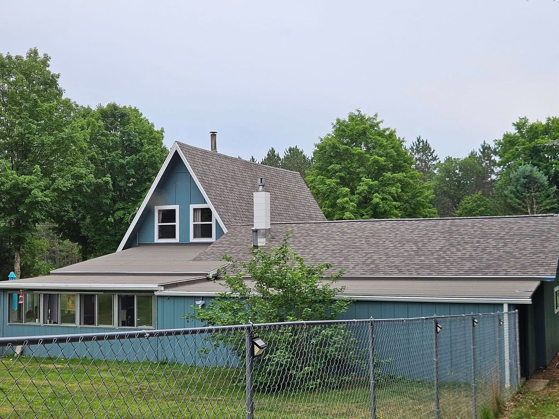 A blue A-frame house with a sprawling attached roof stands behind a chain-link fence surrounded by green trees.
