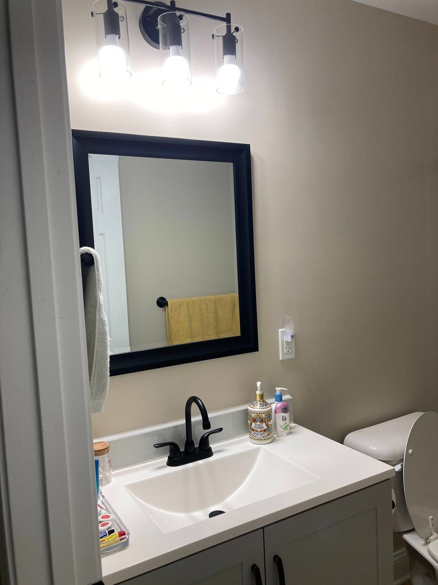 A modern bathroom vanity with a white countertop, black faucet, rectangular black-framed mirror, and three-bulb fixture.