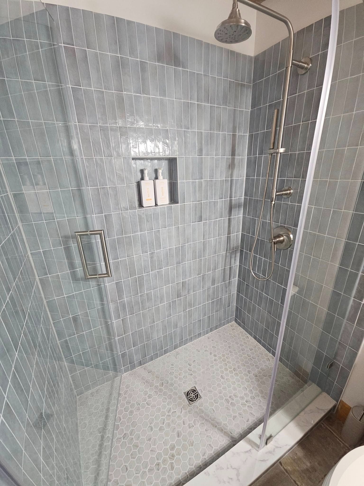 A modern glass-enclosed shower with gray rectangular wall tiles, a rainfall showerhead, and a pebble-stone floor.