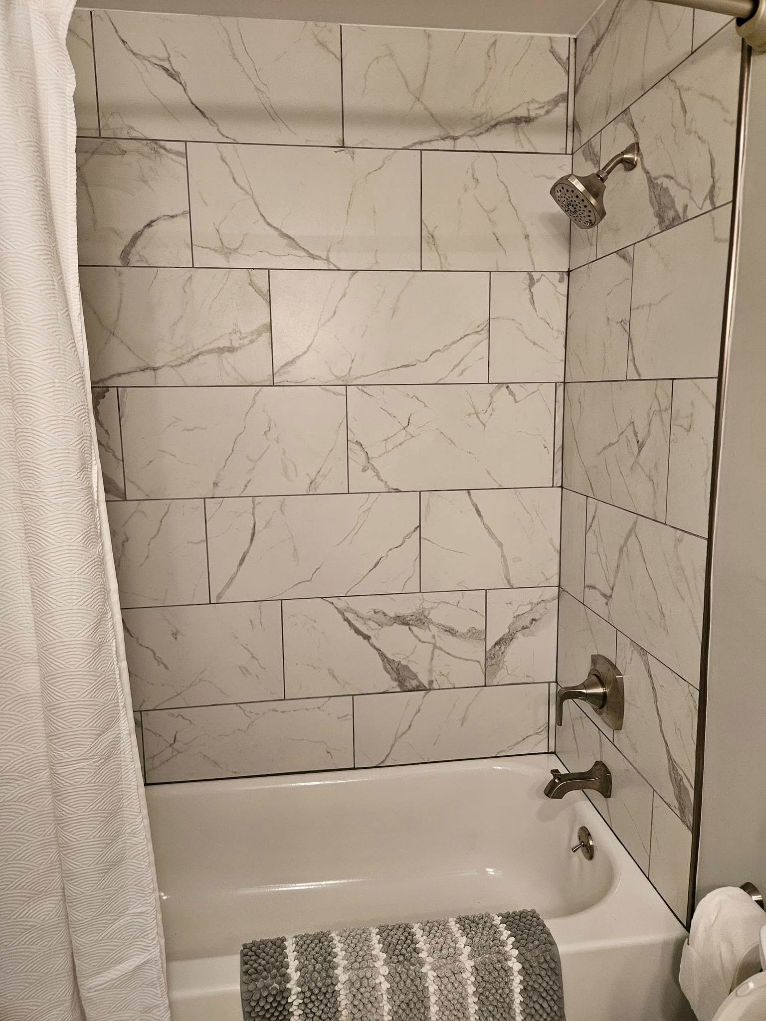 A bathroom shower featuring white marble-patterned tiles, a white bathtub, and silver fixtures.