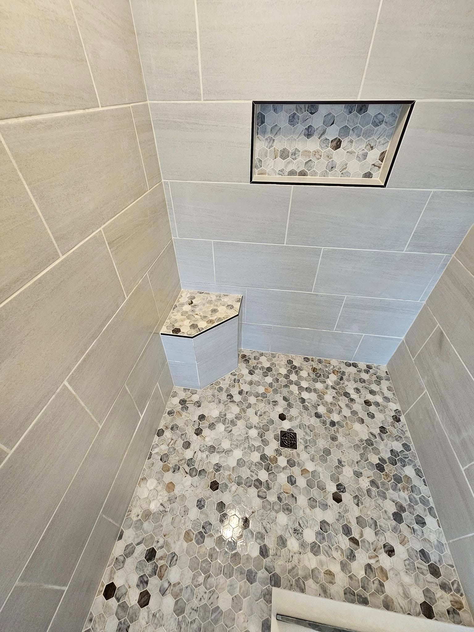 A shower stall featuring large light grey rectangular tiles, a pebble floor, a corner seat, and a tiled wall niche.