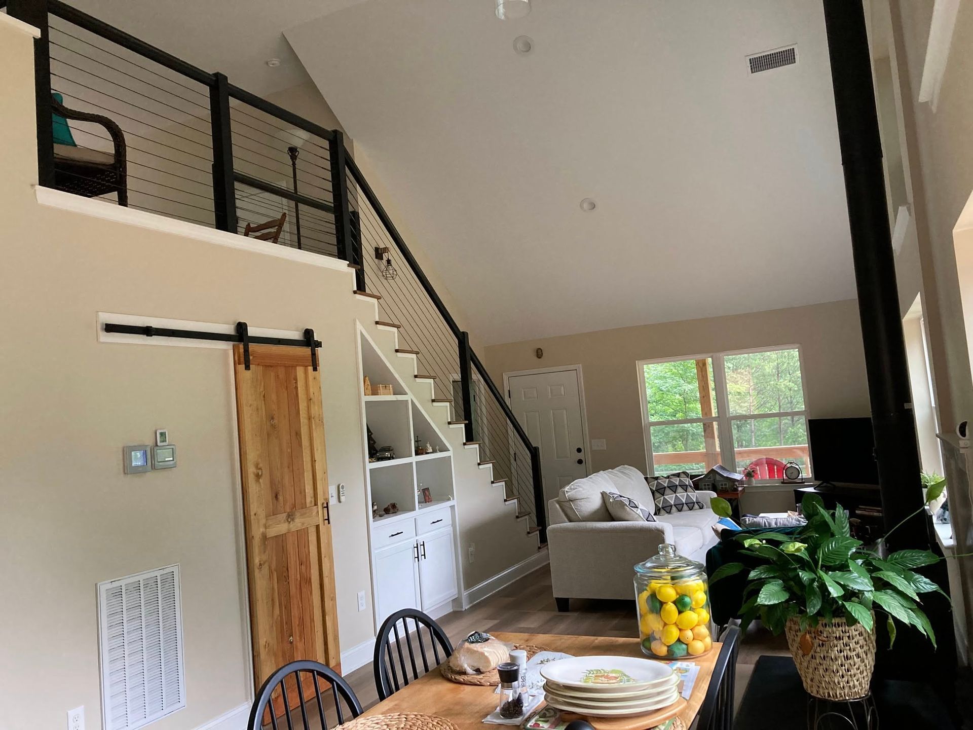 Modern living area with a loft, cable-railed staircase, wooden barn door, dining table, and a living room with a sofa.