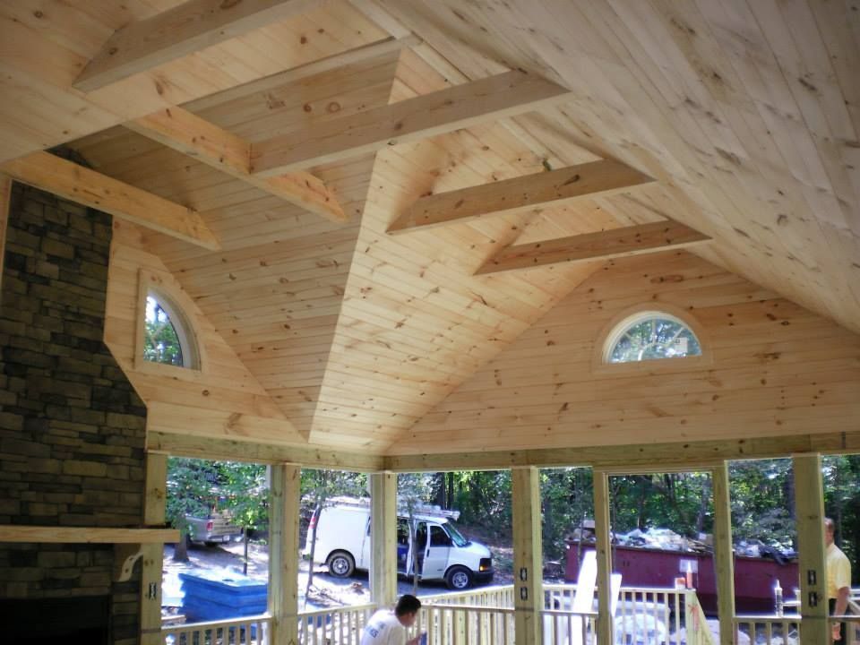 Interior of a screened-in porch with vaulted wood ceilings, exposed beams, a stone fireplace, and two arched windows.