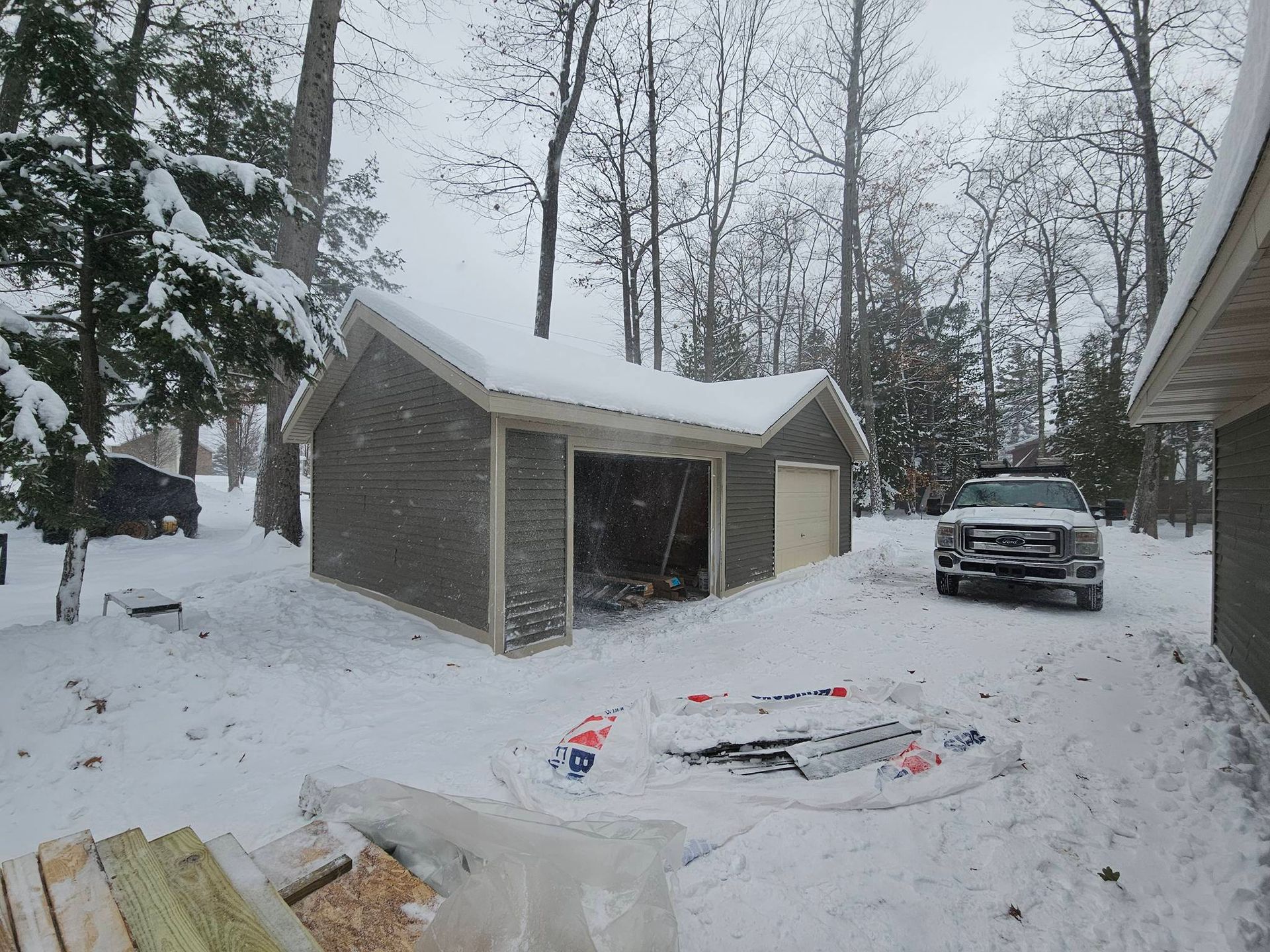 A detached gray garage with a snow-covered roof sits in a snowy, wooded yard with a pickup truck parked nearby.