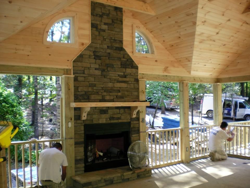 Two workers finish a stone fireplace on a wood-paneled porch with arched windows and a view of trees and a van.