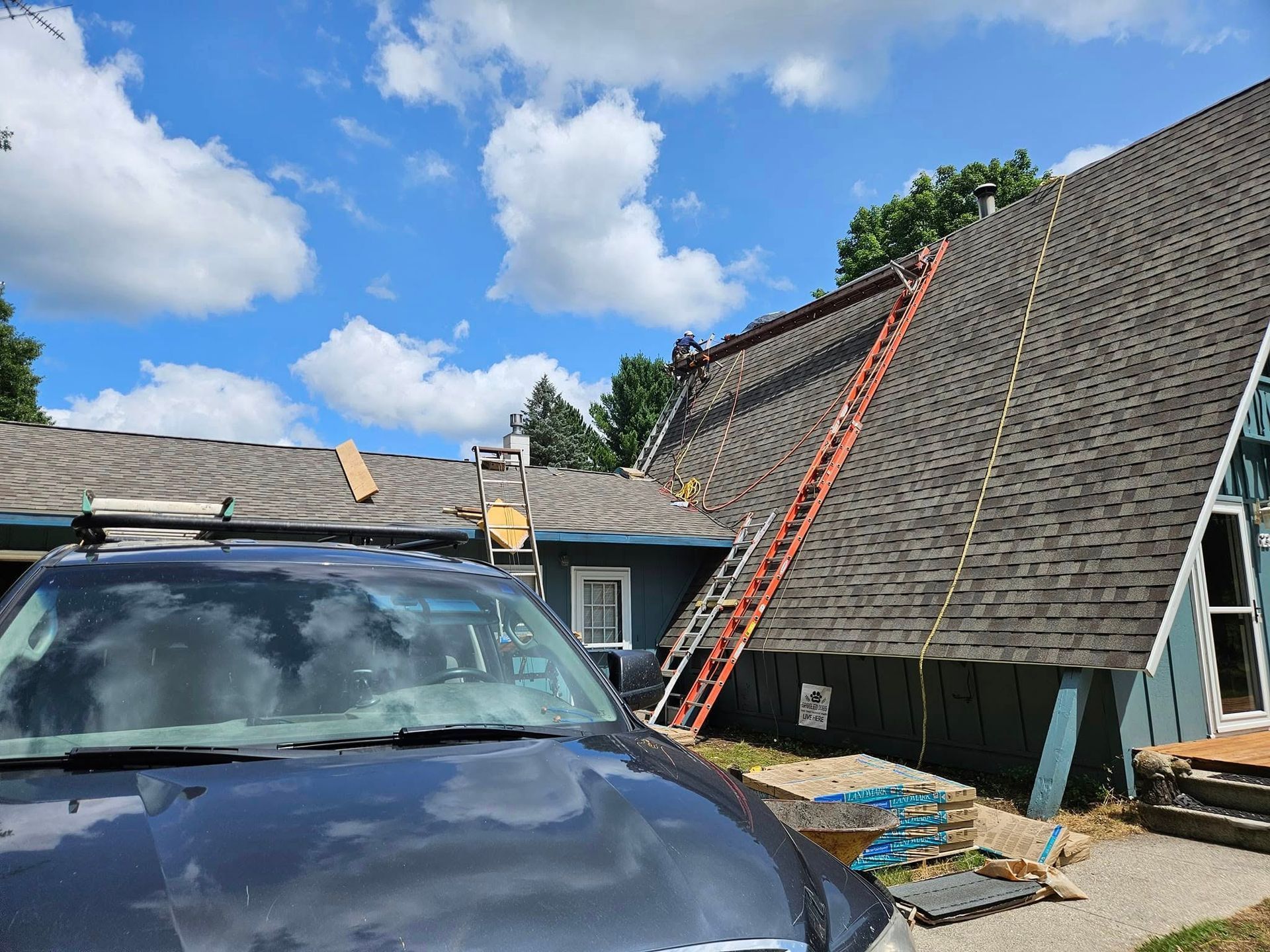 A dark-shingled A-frame cabin with a wrapped chimney, exterior support beams, and a small attached section under blue skies.
