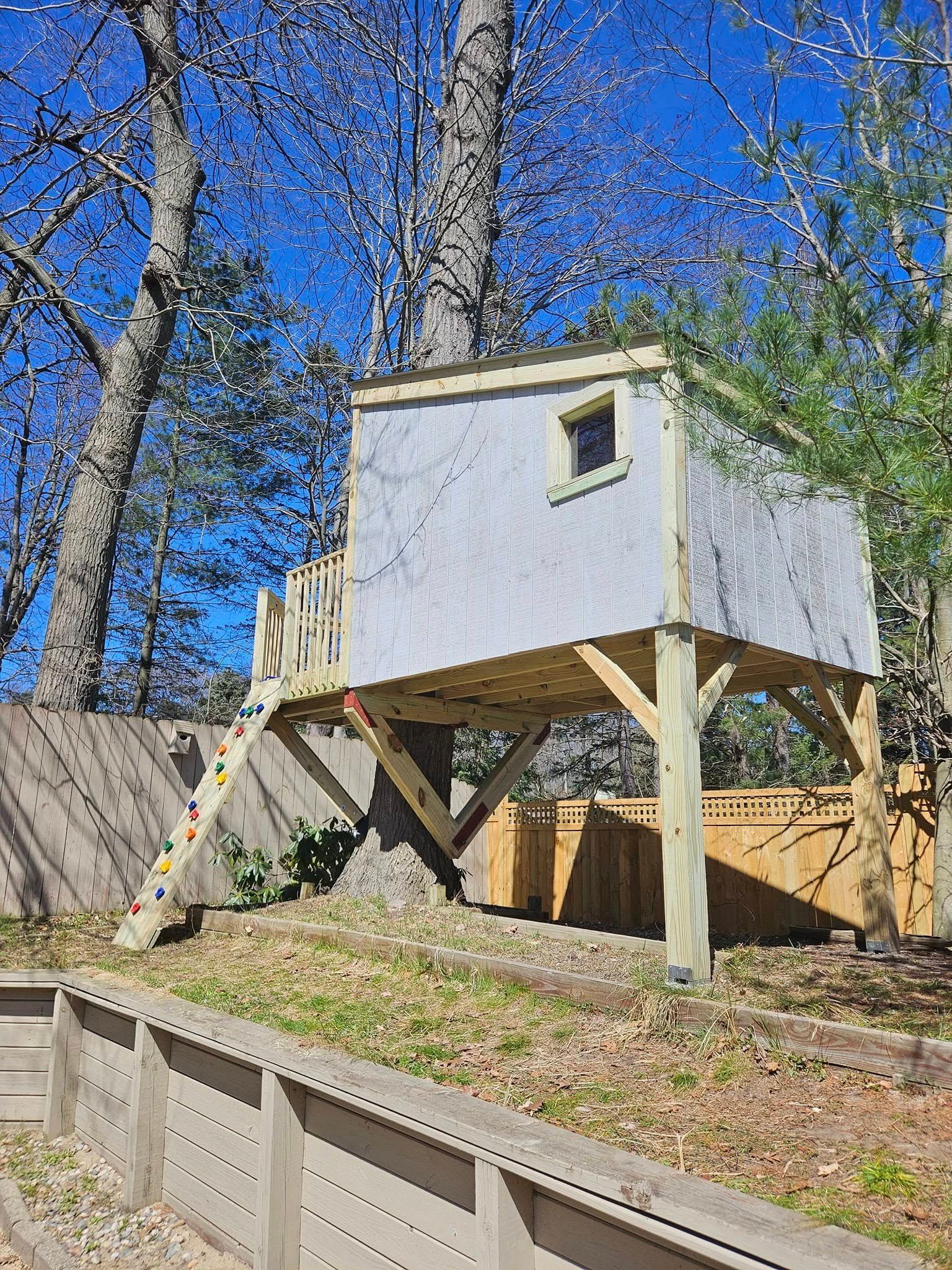 A grey wooden treehouse with a climbing wall, supported by posts in a backyard with a retaining wall and trees.