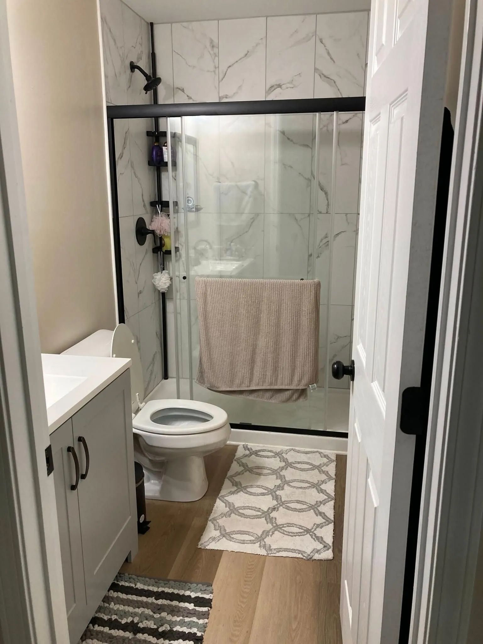 A small bathroom with a gray vanity, a toilet, a shower with marble-patterned tile, and patterned floor rugs.