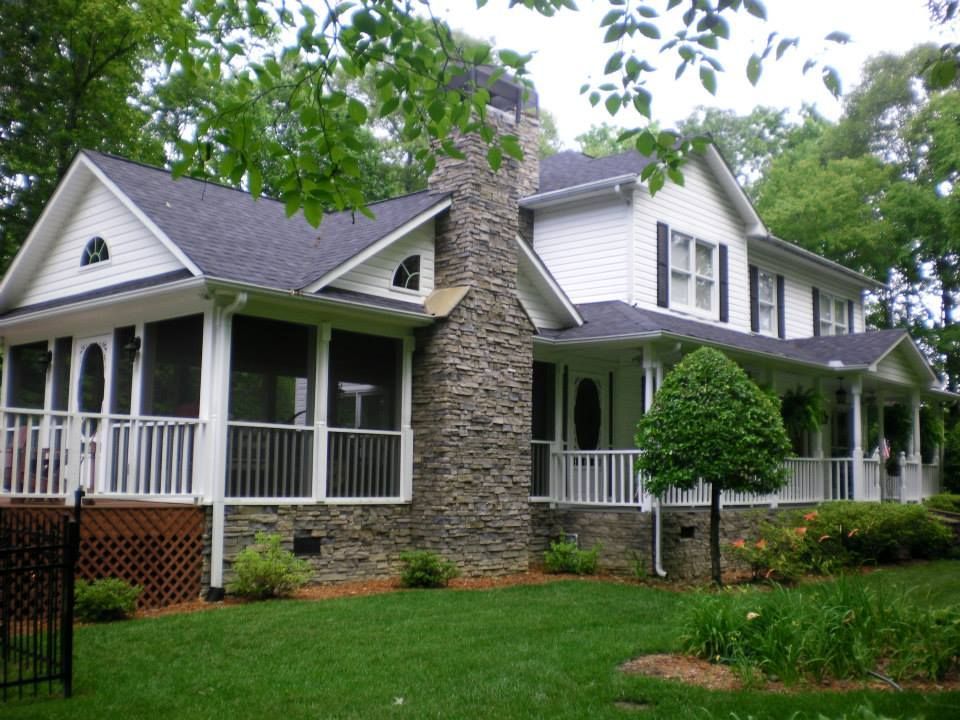 A white, two-story house with a large stone chimney, wrap-around porch, and screen enclosure, set in a green yard.