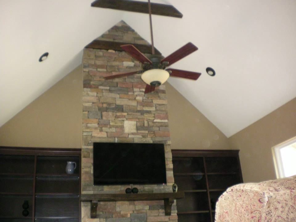 A stone fireplace with a mounted television and dark wood shelves, set against a vaulted ceiling with a ceiling fan.