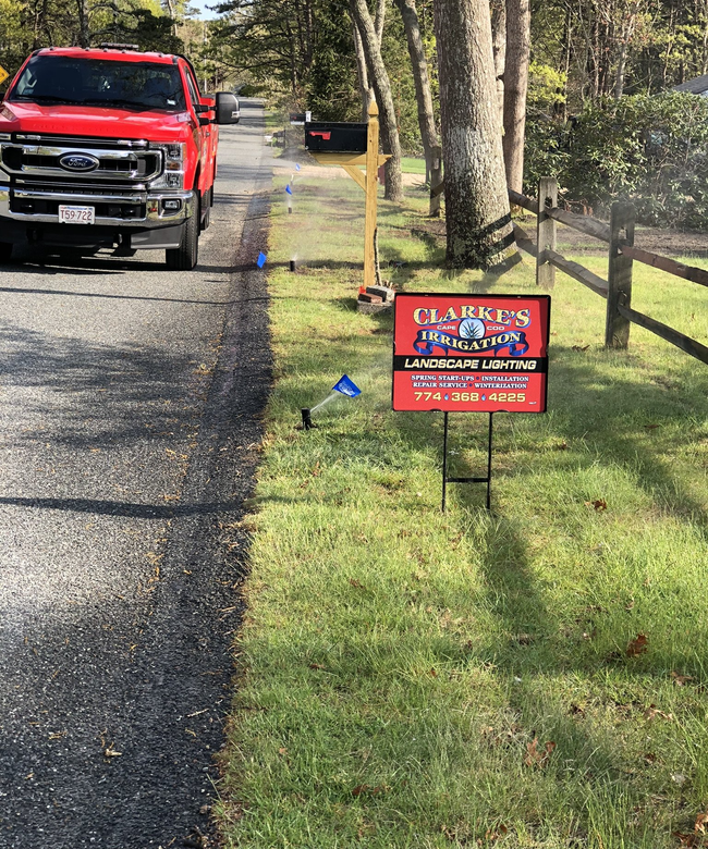 A red Ford truck parked next to a lawn with a Clark's Landscaping business sign and utility marking flags.