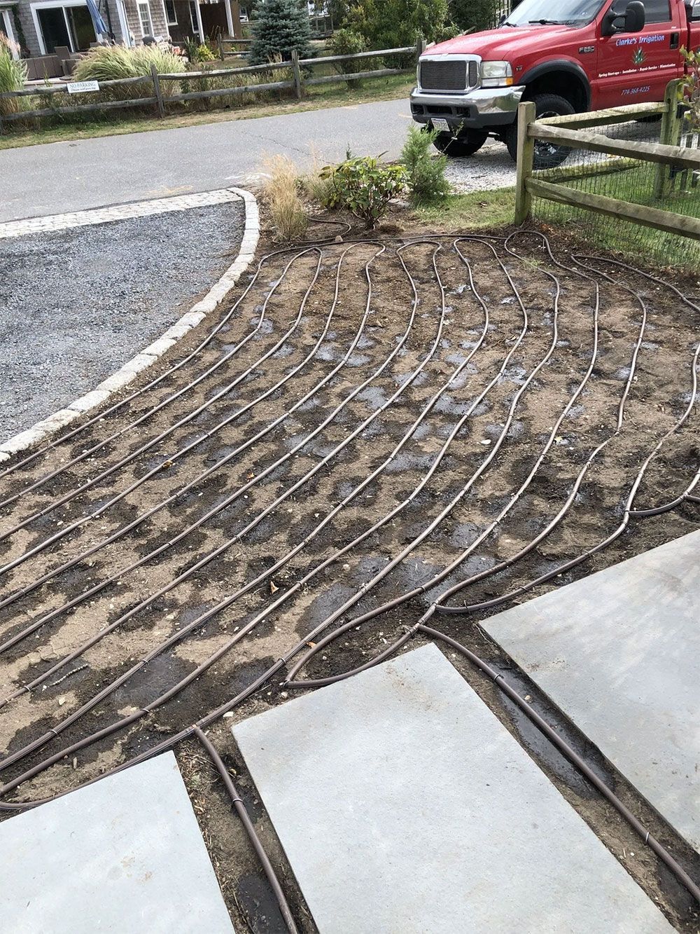 Black irrigation tubing laid in parallel lines across a soil garden bed, positioned next to stone pavers and a driveway.
