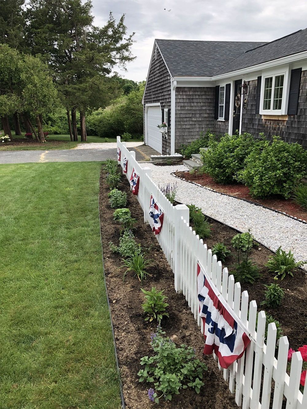 A white picket fence decorated with patriotic bunting lines a mulched garden bed in front of a gray shingled house.