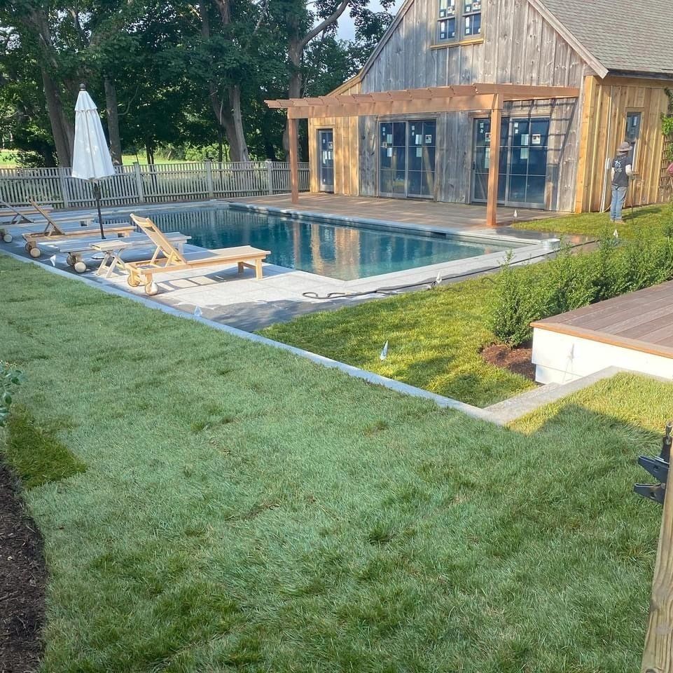 A rectangular swimming pool with lounge chairs and a white umbrella sits in front of a rustic wooden barn-style building.