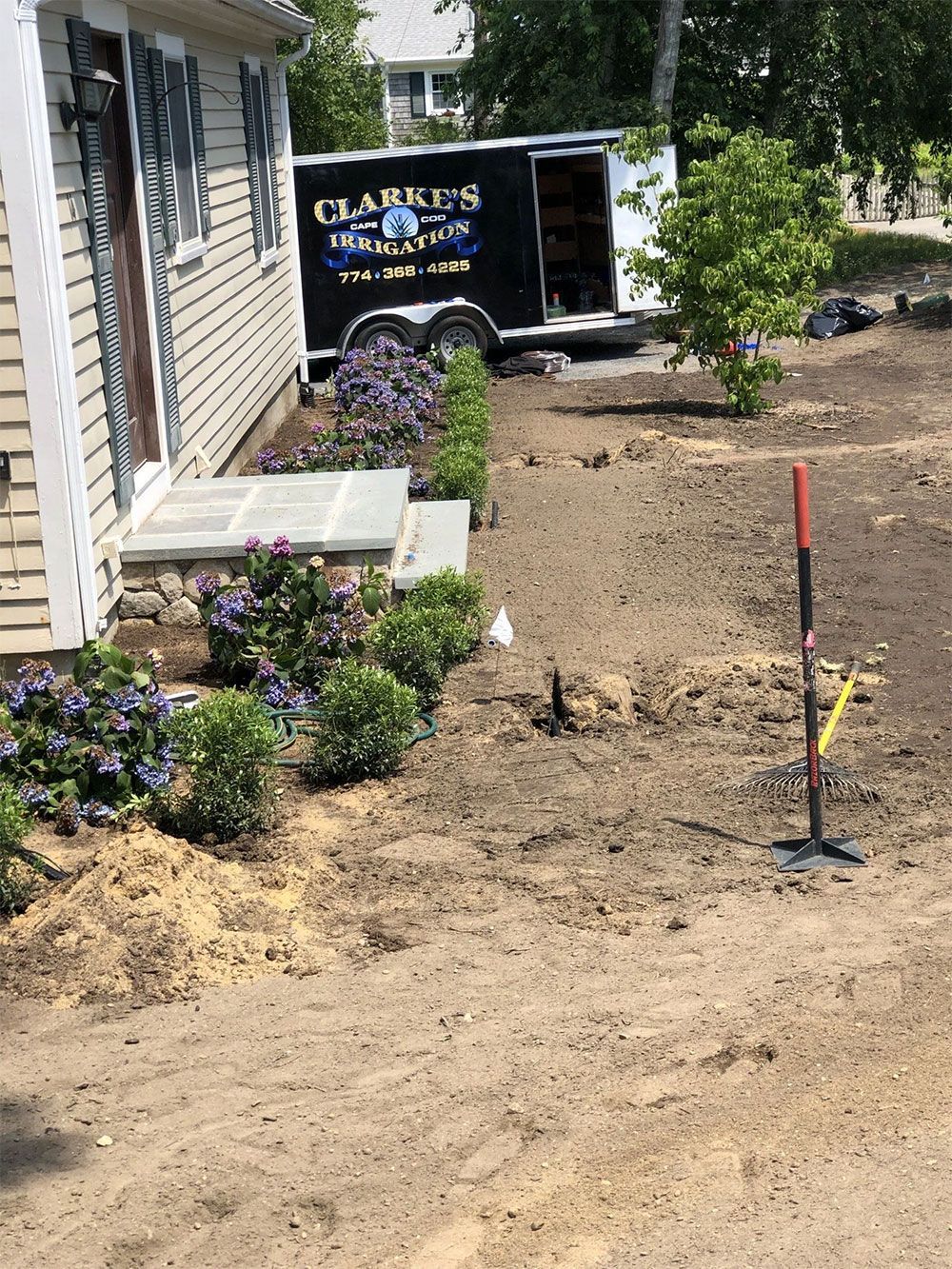 A residential yard undergoing landscaping with freshly planted shrubs and a contractor trailer parked in the background.