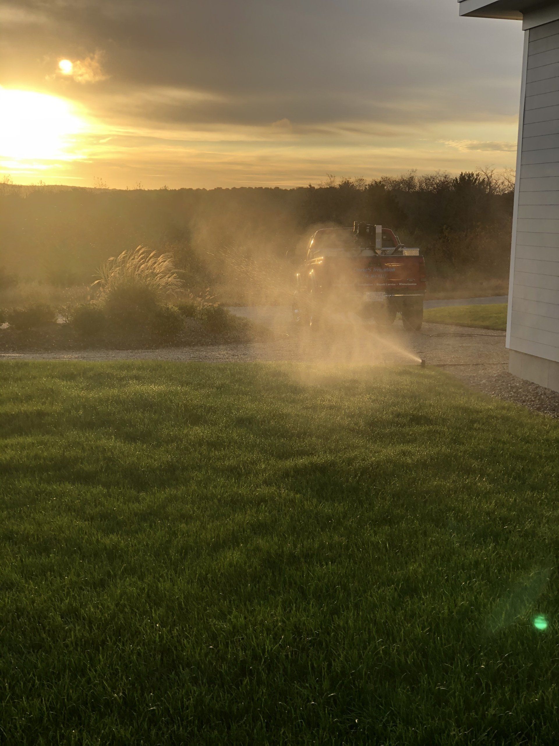 A lawn sprinkler sprays a fine mist in a sunlit yard at sunset, near the corner of a white building.