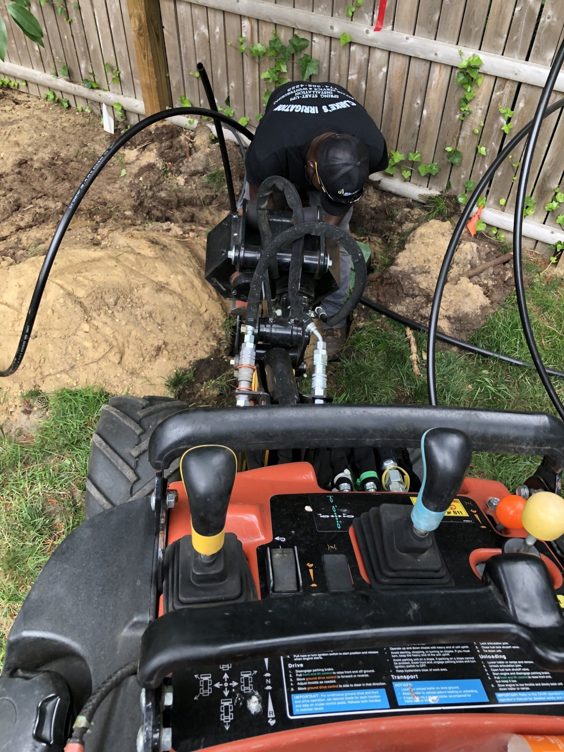 A person operates a trencher in a yard, digging a path along a fence line.