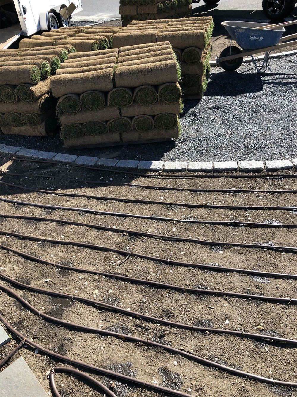 Stacks of rolled sod sit on a gravel driveway near a yard prepared with black drip irrigation tubing for installation.