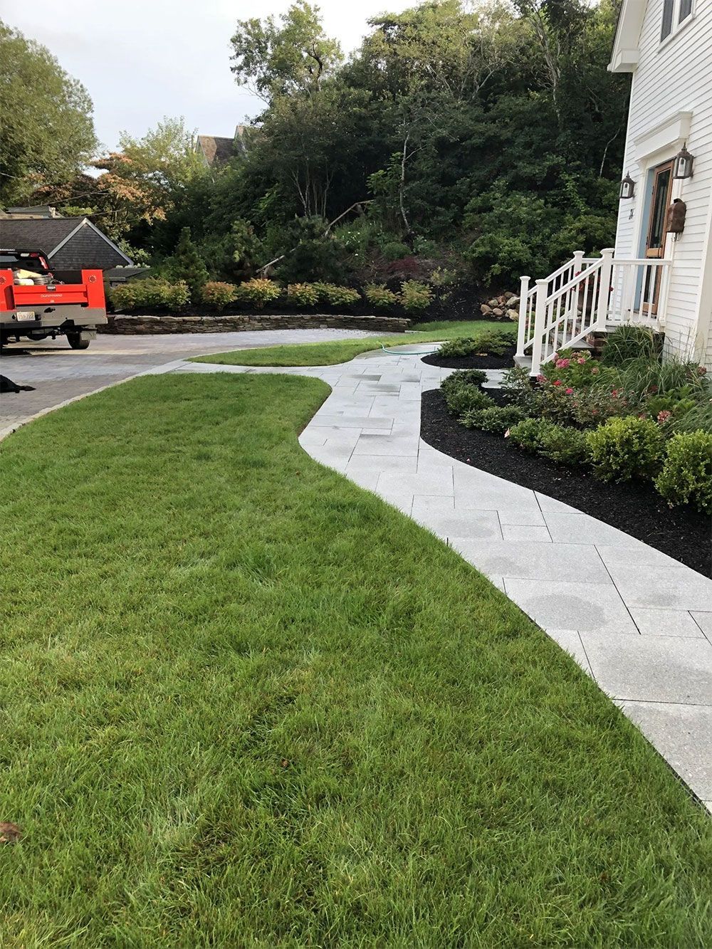 A winding paved stone path leads to the front porch of a white house with a green lawn and dark mulch landscaping.