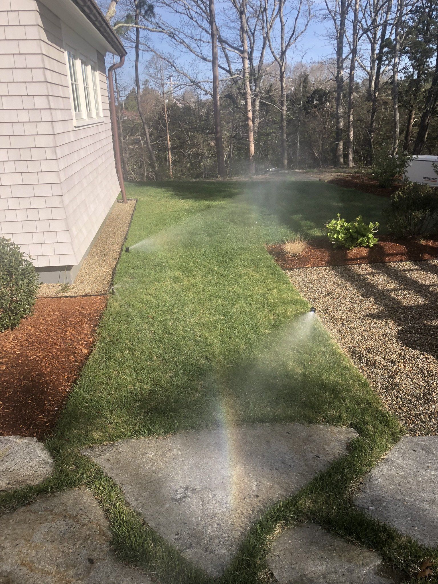 A lawn sprinkler waters a grassy yard between a house, garden beds, and stone pavers, creating a small rainbow.