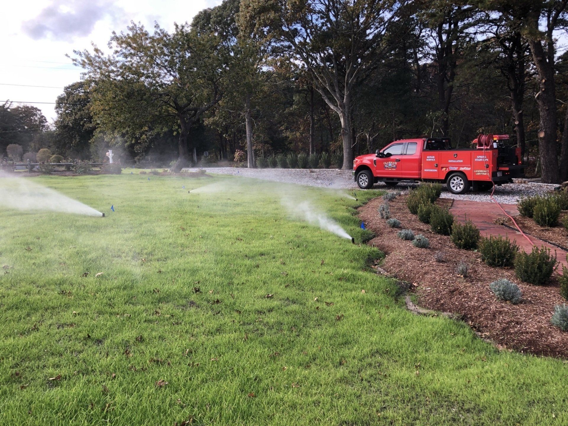 A bright red utility truck parked near a landscaped bed while lawn sprinklers water a green yard.