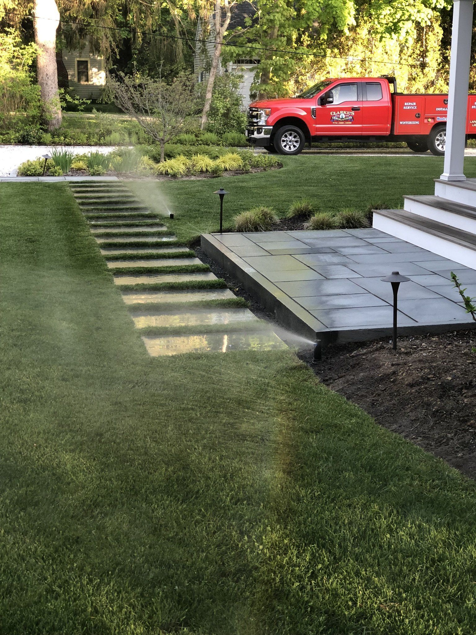 A sprinkler waters a lawn next to a stone walkway and porch with a red utility truck parked in the background.