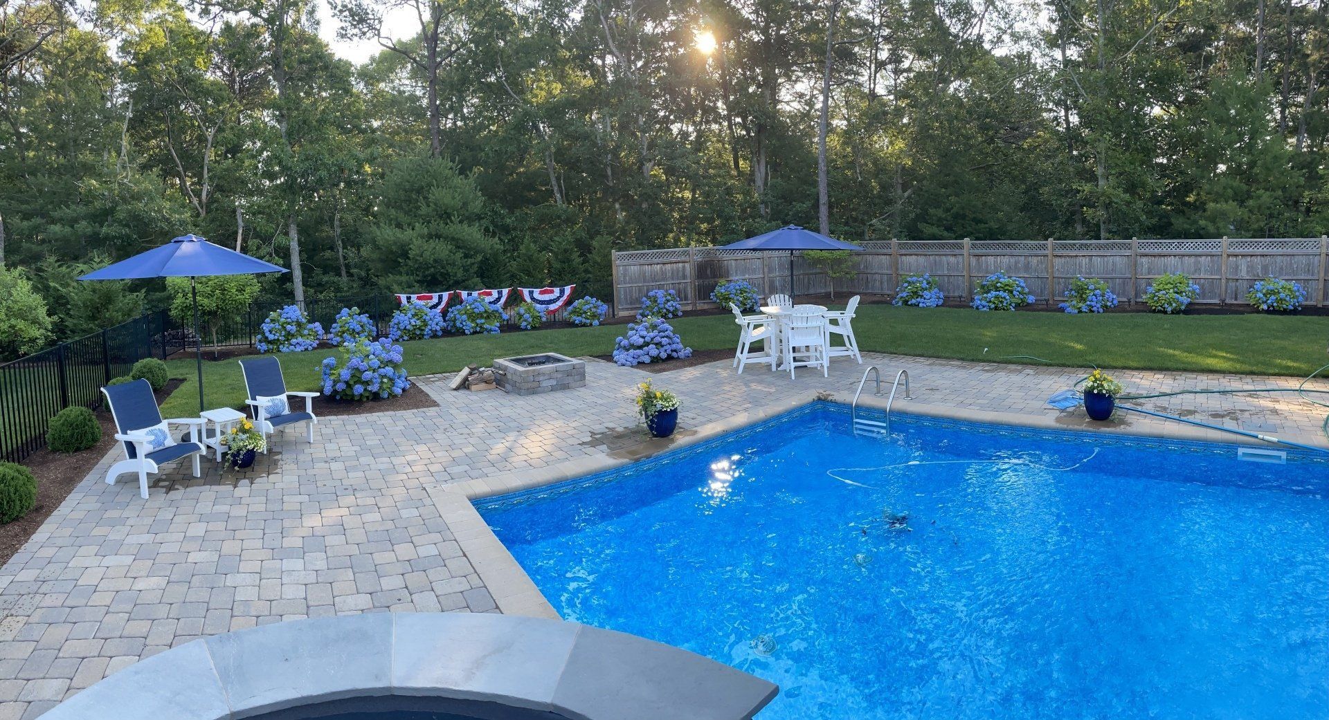 A sunny outdoor backyard with a blue swimming pool, stone patio, blue patio umbrellas, and lush landscaping.