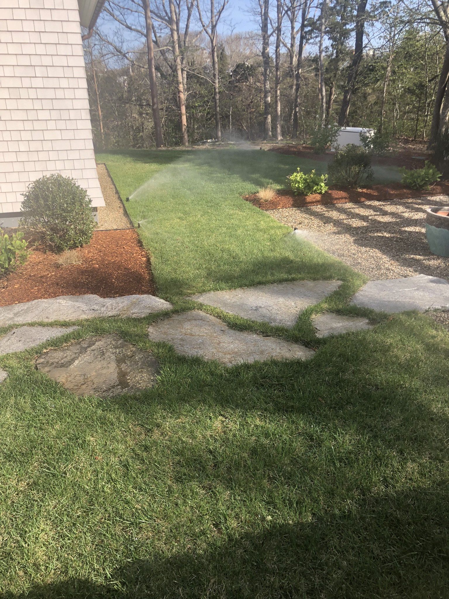 Stone stepping stones embedded in a grassy lawn next to a building wall with a sprinkler spraying in the background.