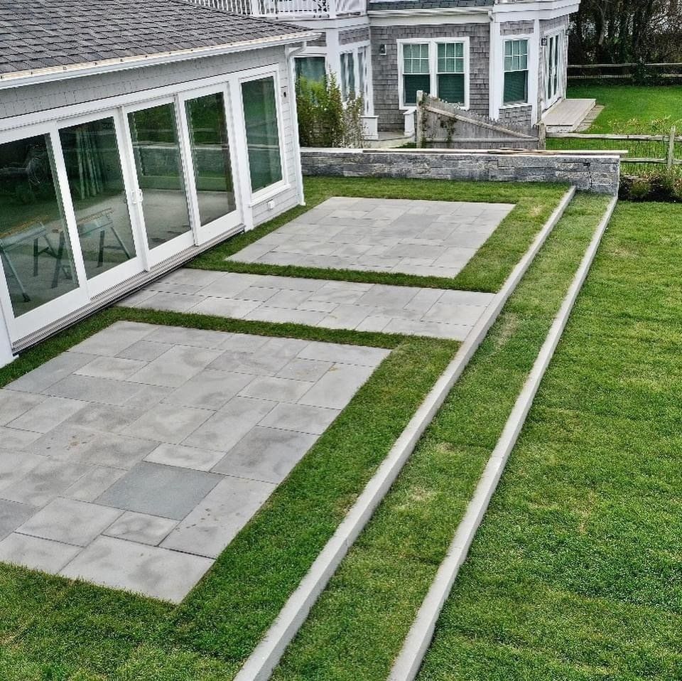 A modern home patio with two grey paver sections separated by grass, featuring a long stone step leading to a lawn.
