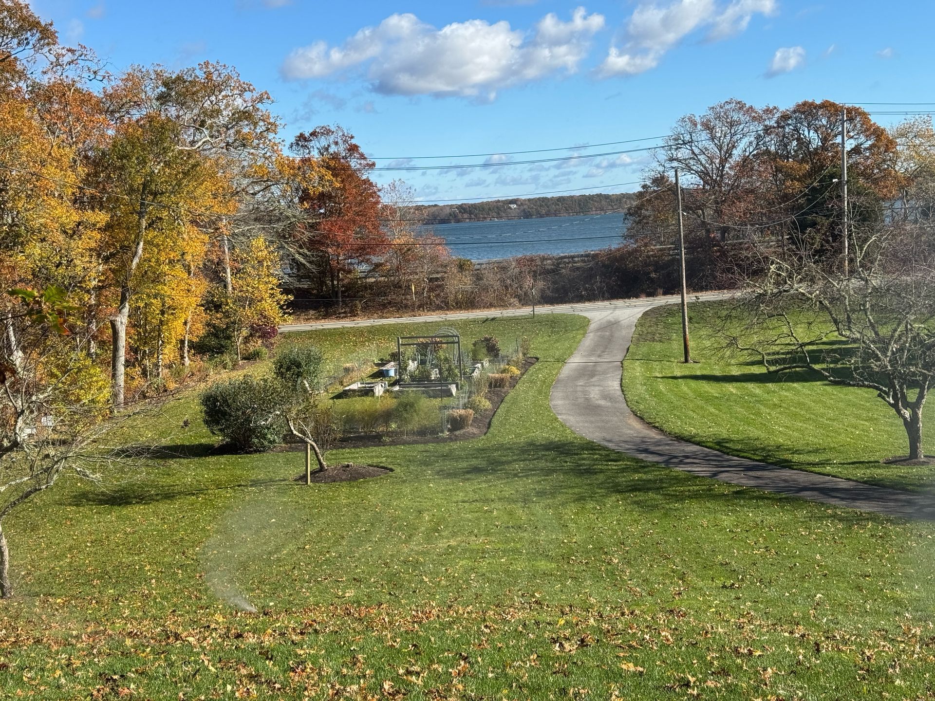 A grassy lawn with a garden bed and a curving path leading toward a distant body of water under a blue, cloudy sky.