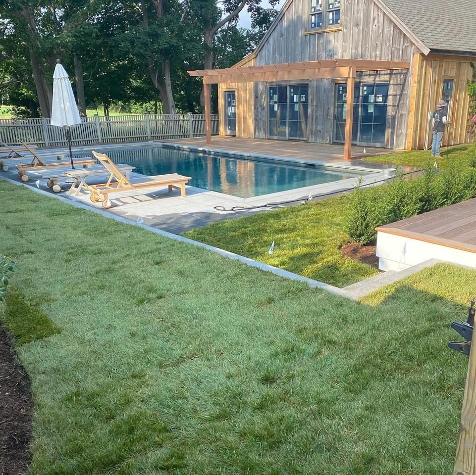 A rectangular swimming pool sits next to a rustic wooden barn with a pergola and lounge chairs on a grassy lawn.