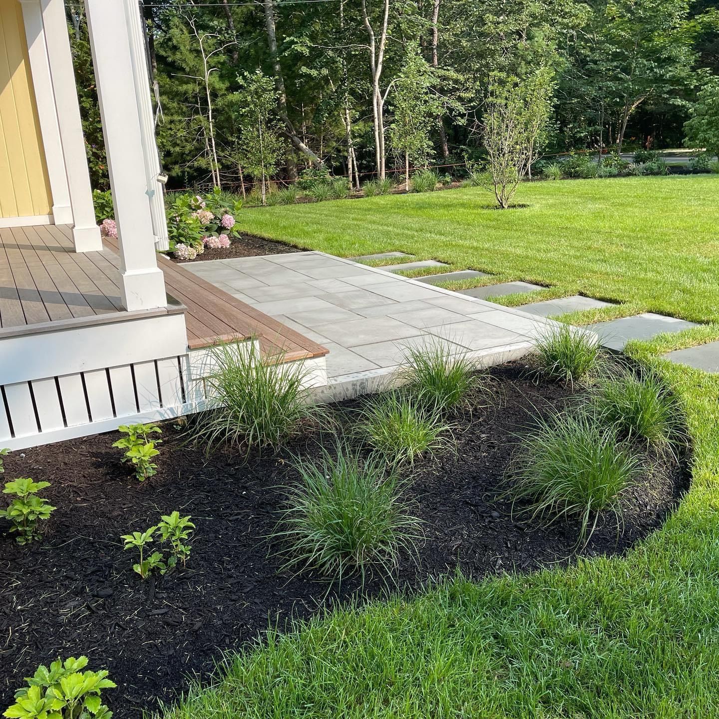 A light yellow house porch connects to a gray stone patio with a path of stepping stones leading into a green lawn.