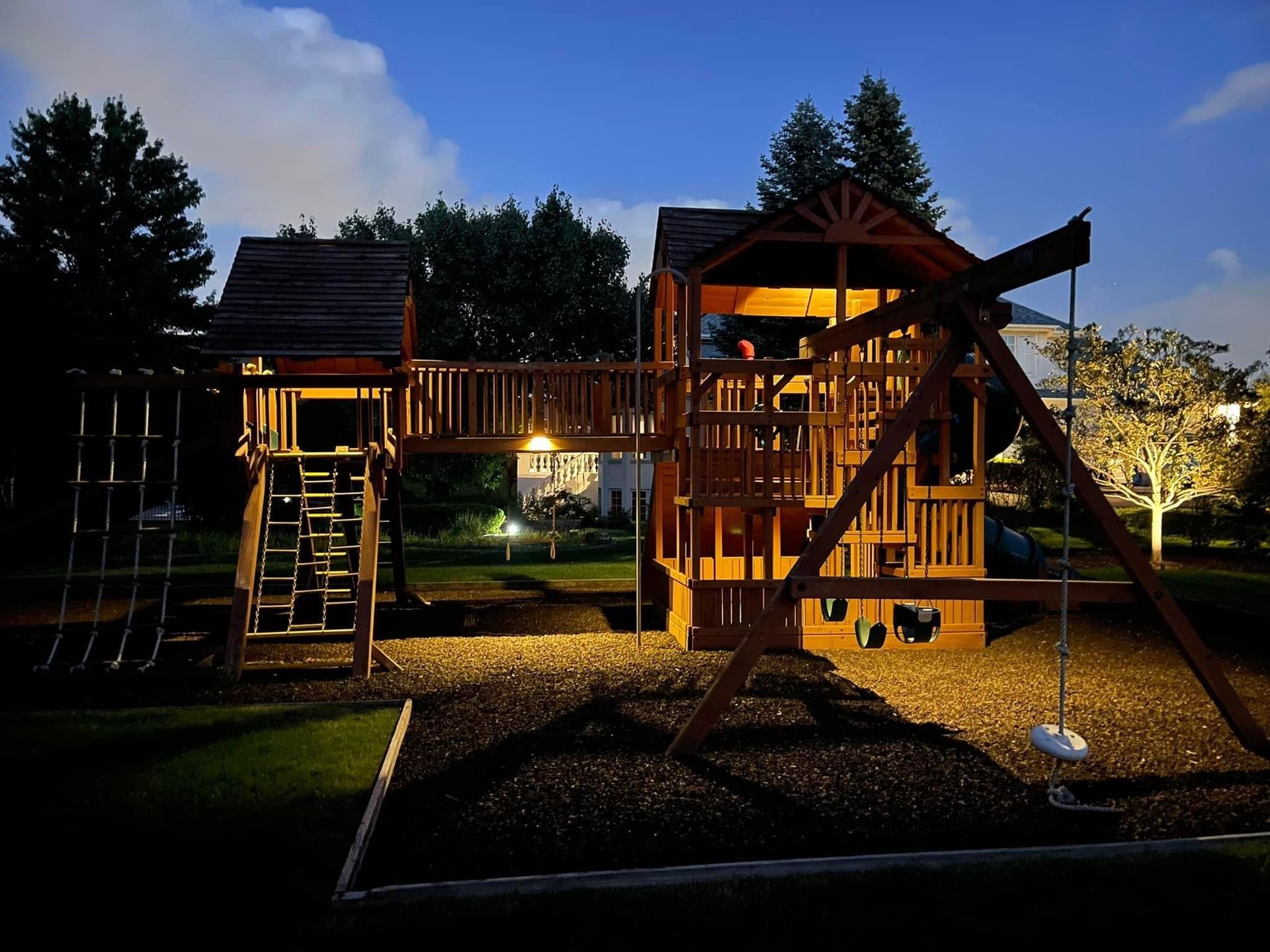 A large wooden backyard playground with a swing set and climbing structures illuminated at night.