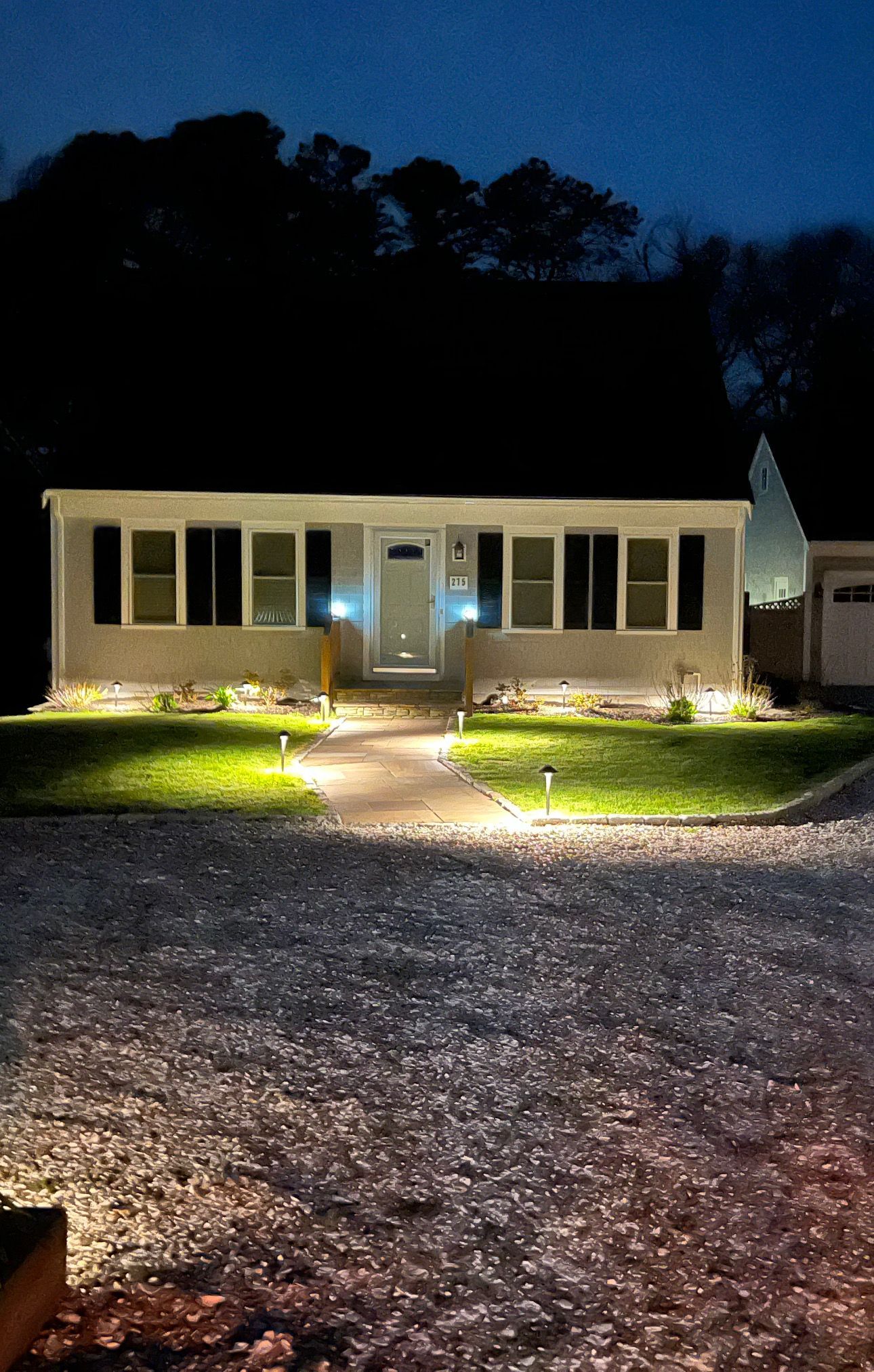 A single-story gray house at night, illuminated by warm outdoor landscape lighting and a bright front porch light.