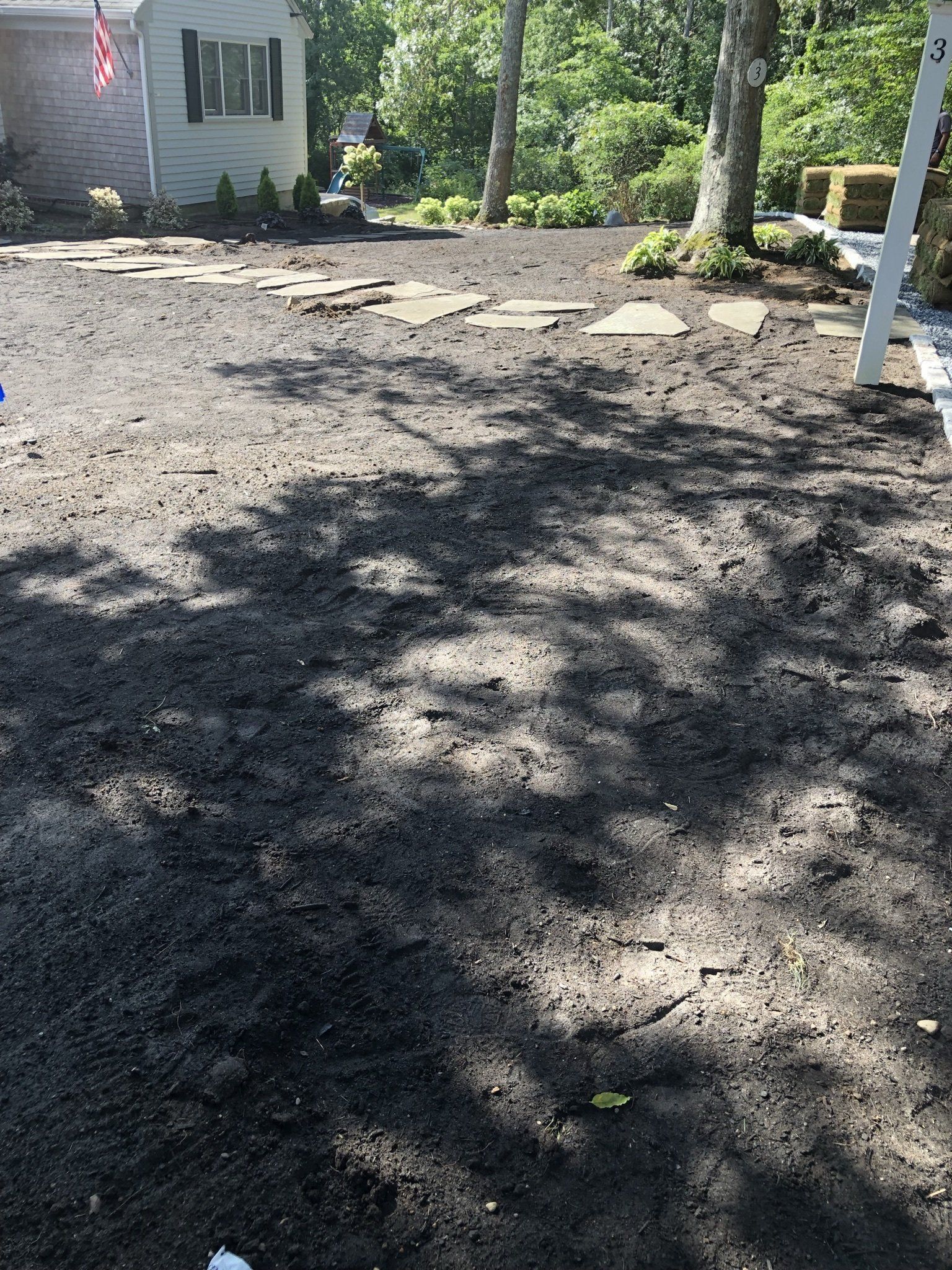 A yard with fresh dark soil, a stone walkway leading to a house, and mature trees casting shadows.