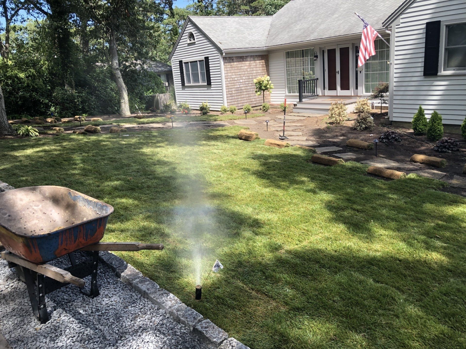 A wheelbarrow sits on gravel next to a green lawn being watered by an upright sprinkler in front of a house.