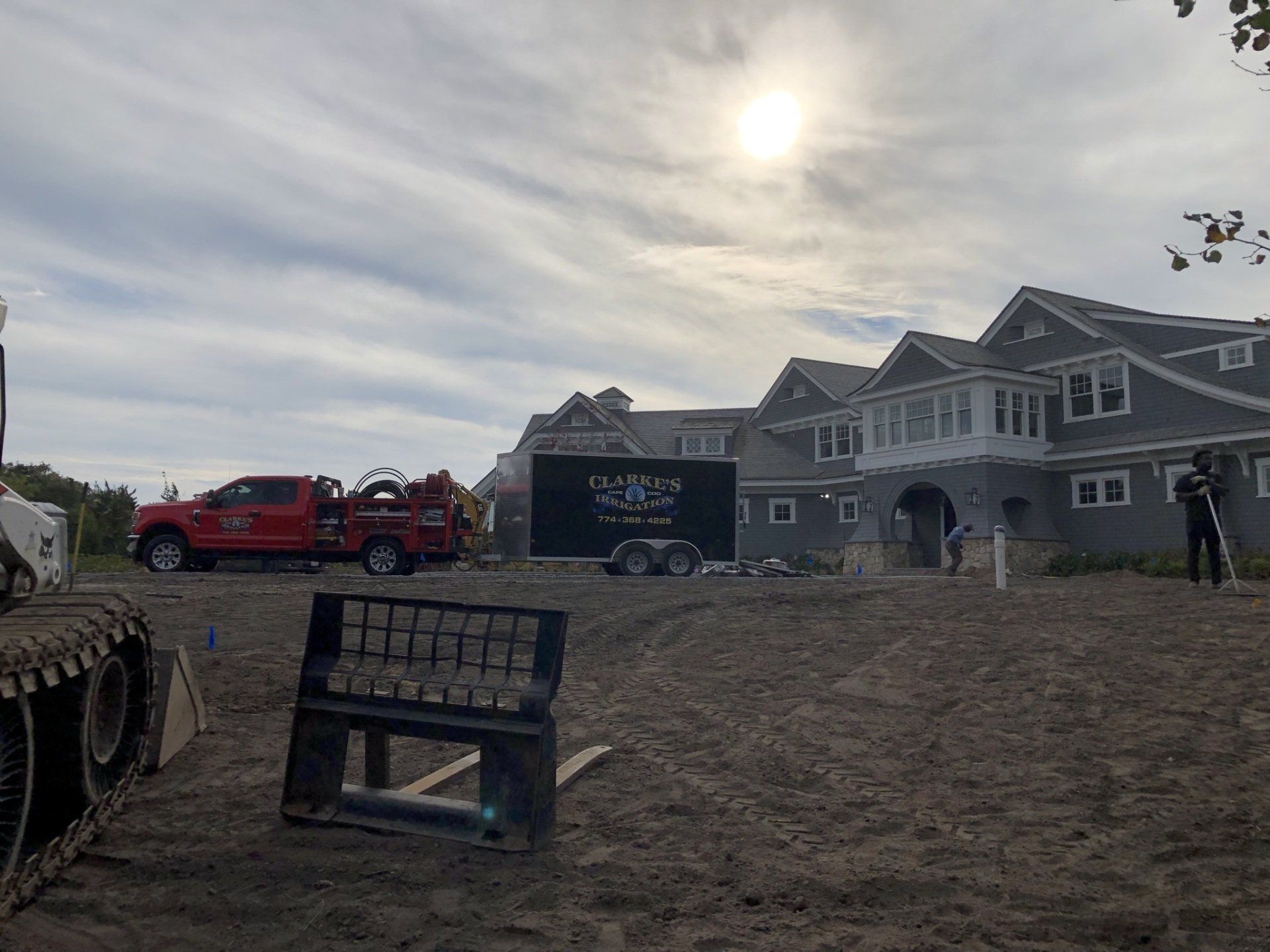 A red utility truck and construction equipment sit on a dirt lot in front of a large, multi-story grey house.
