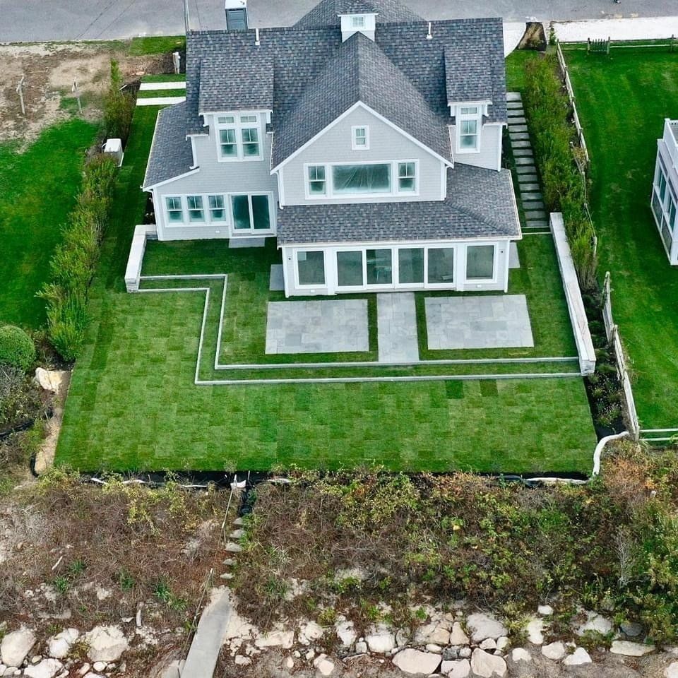 An aerial view of a light gray, multi-story coastal home with a fresh sod lawn, stone patio, and stone retaining walls.
