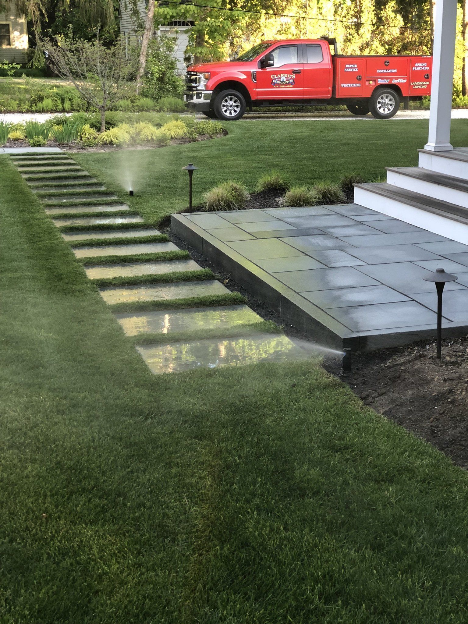 A sprinkler waters a residential lawn next to a modern stone walkway and a gray patio, with a red service truck in the back.