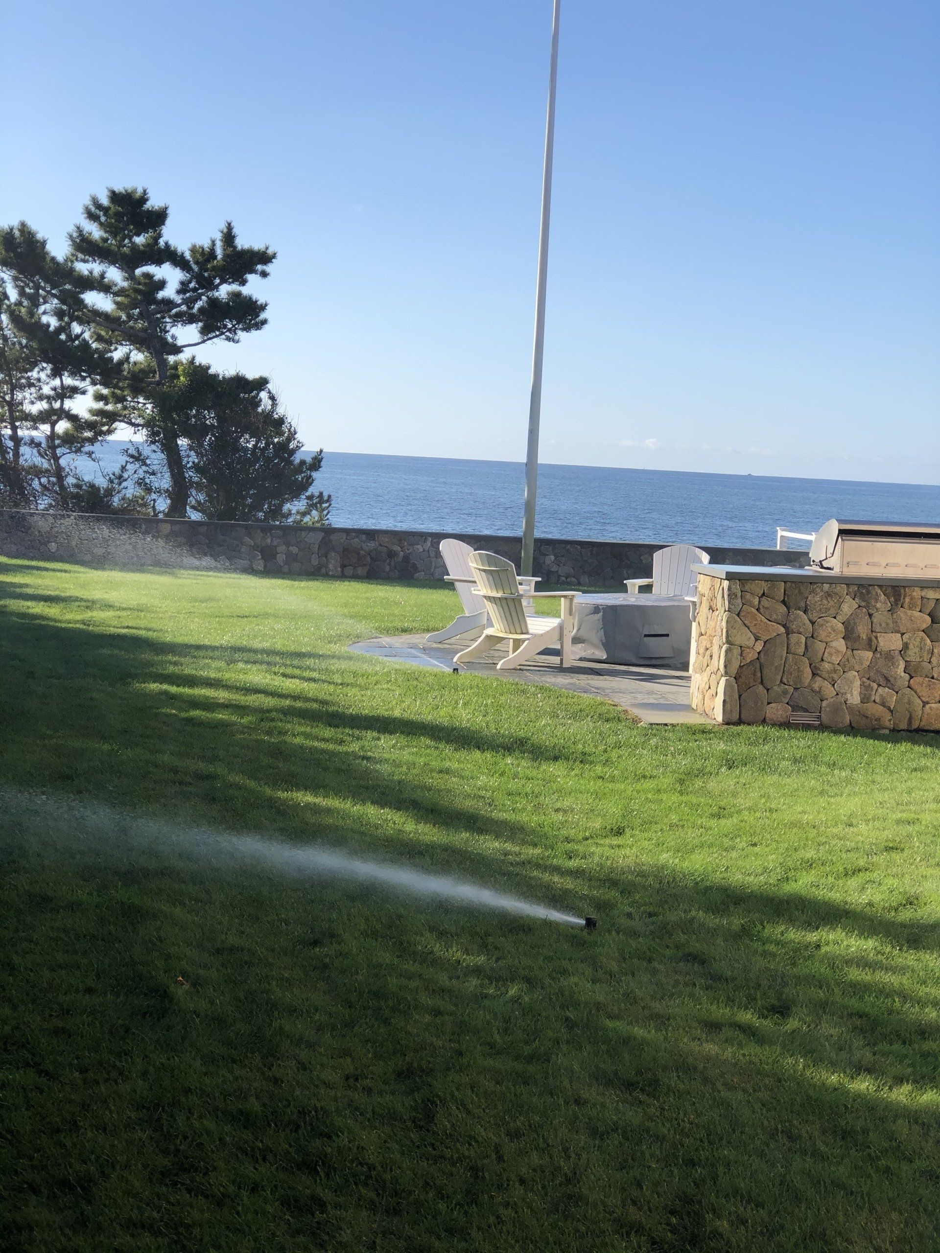 A lawn sprinkler waters a grassy yard by the ocean, with stone walls, chairs, and a fire pit visible in the background.
