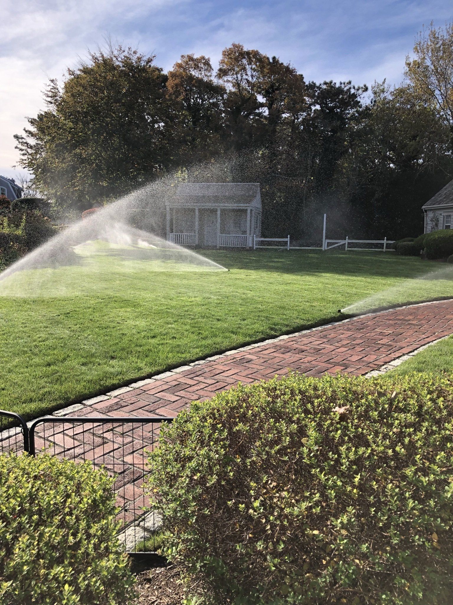 A sprinkler waters a green lawn next to a brick path, with a white structure and trees in the background.