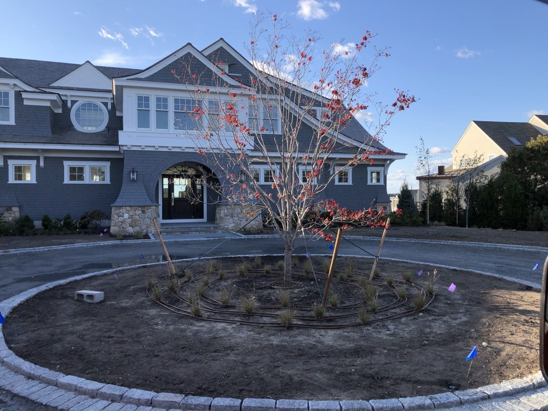 A circular driveway with a young tree in the center leads to a grey house with stone accents under a clear blue sky.