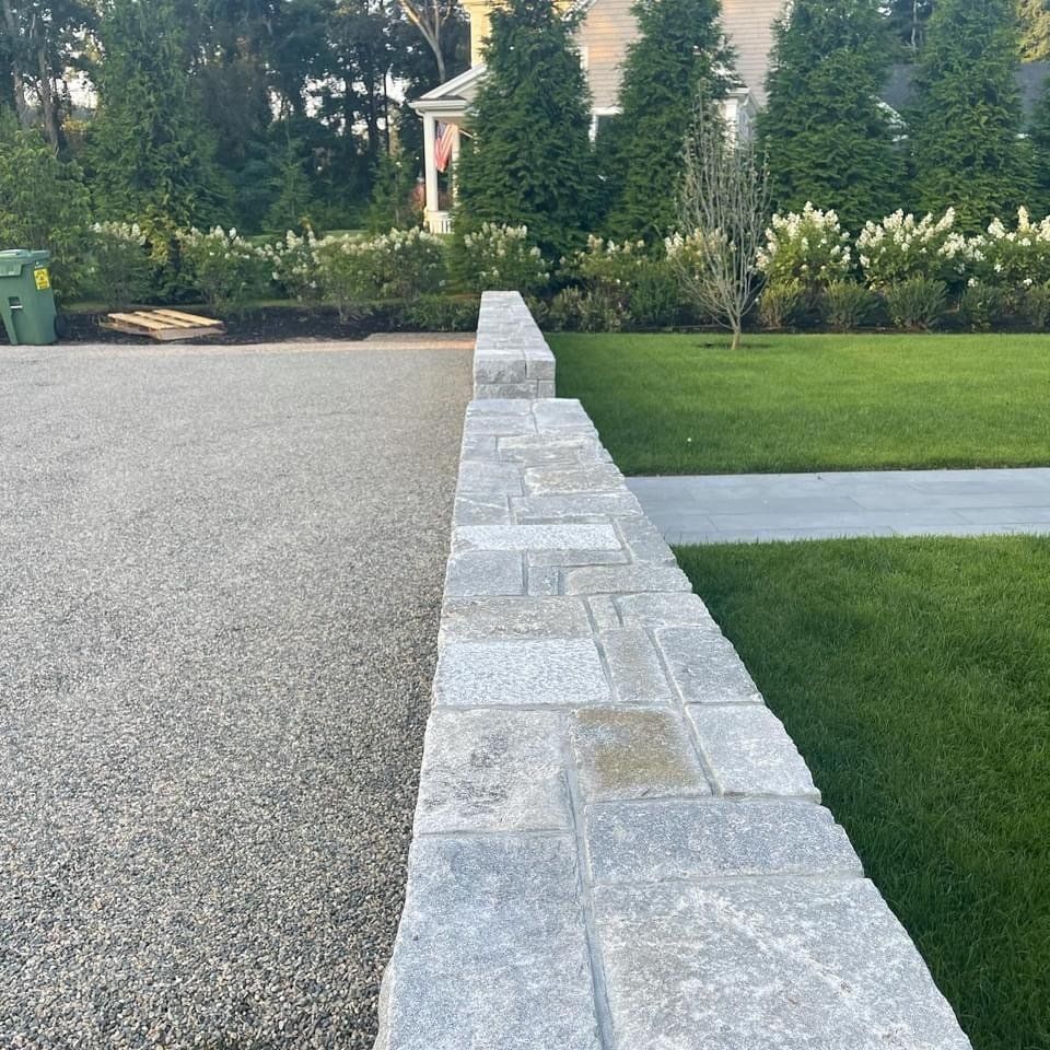 A stone wall separates a grey gravel driveway from a lush green lawn, with a house and manicured trees in the background.
