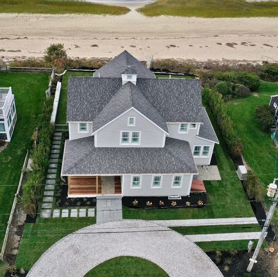 Aerial view of a light gray, multi-roofed coastal home with a wood porch, situated next to a sandy beach.