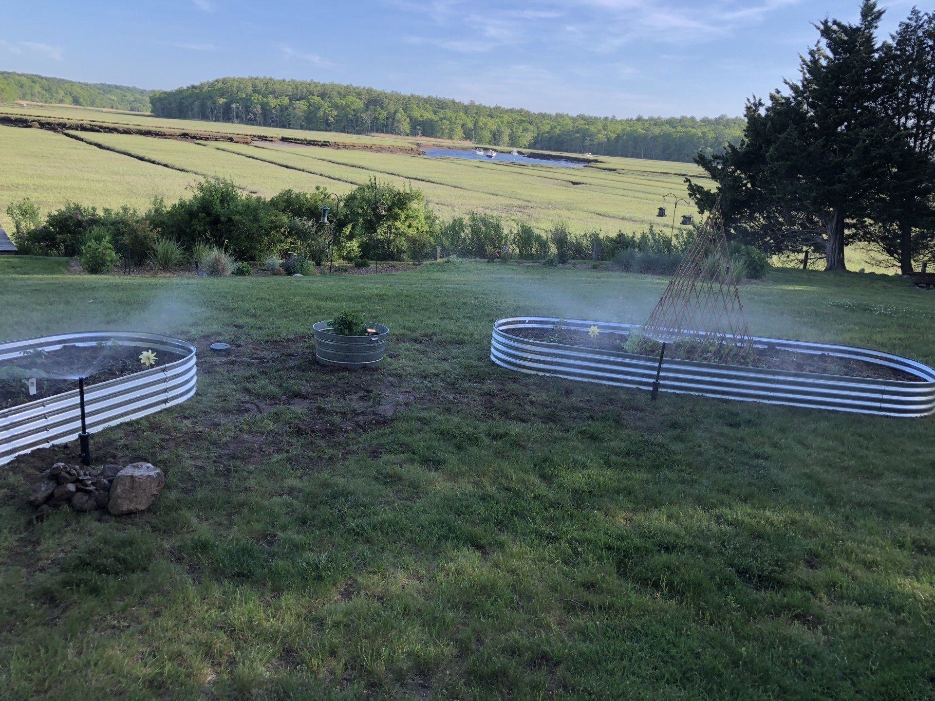Two corrugated metal raised garden beds sit in a grassy field, being watered by misting irrigation sprayers.