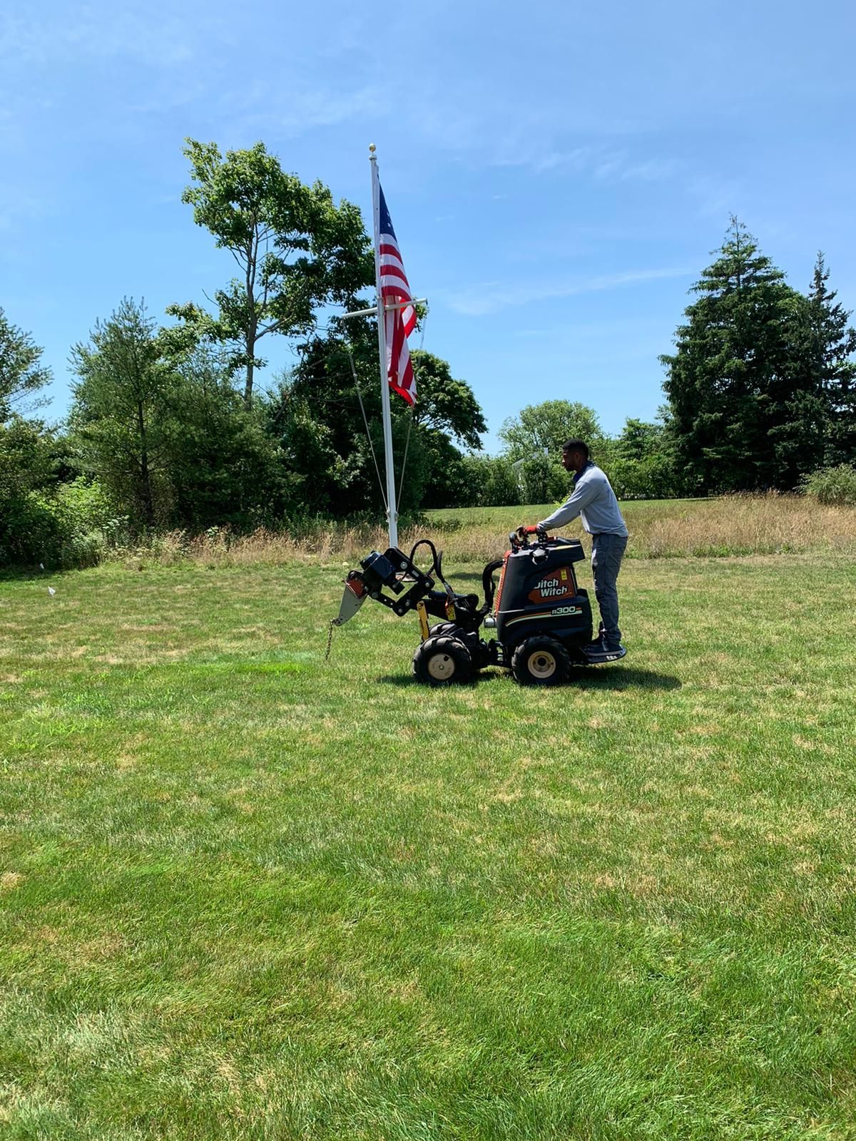 A person operating a stump grinder in a grassy field beneath an American flag on a sunny day.