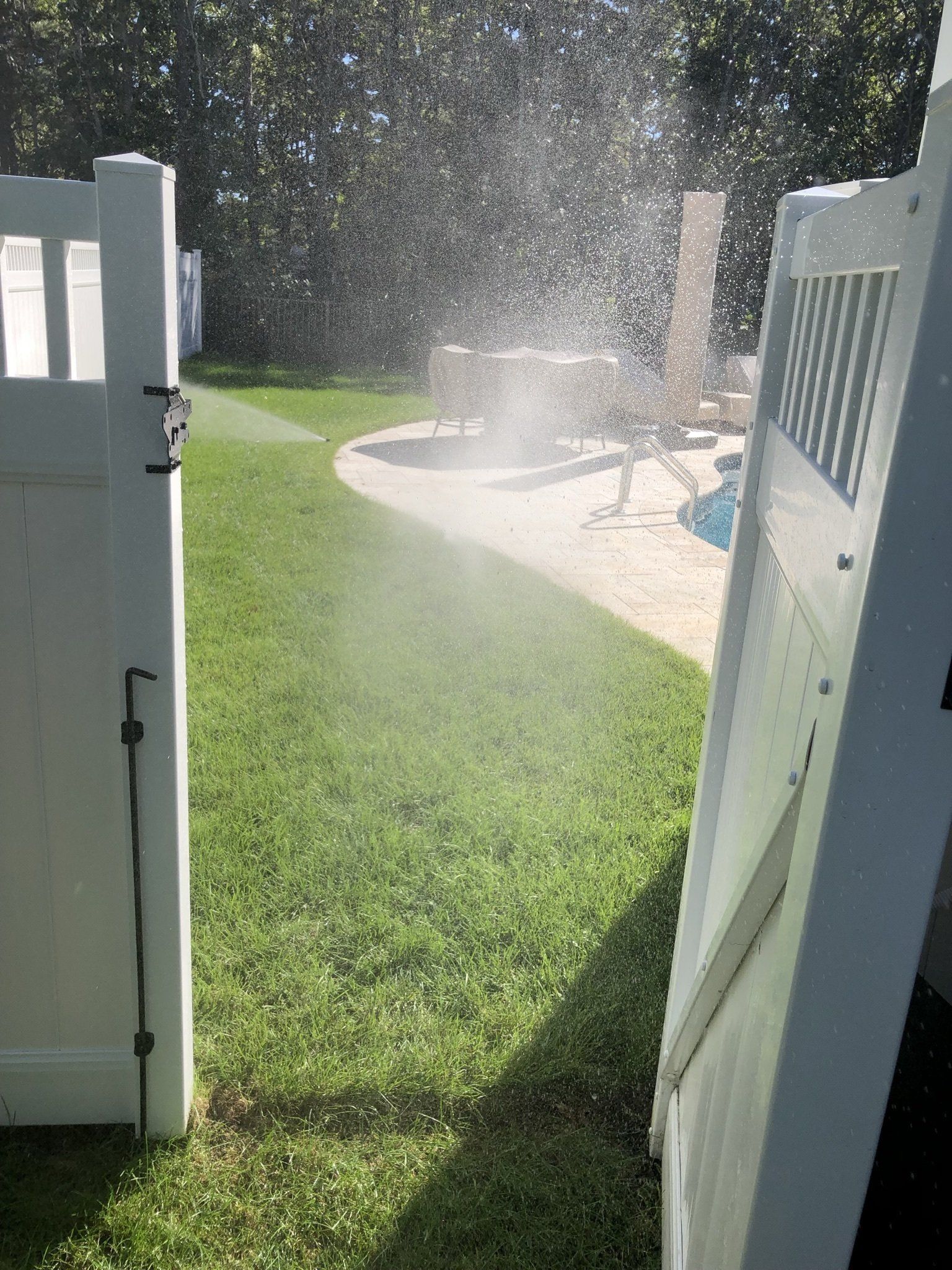 A view through a white gate opening showing a backyard with grass, a pool area, and a fine mist of water in the sunlight.